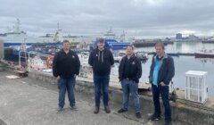 Four people stand on a dock in front of several docked boats and ships under a cloudy sky. Industrial buildings are visible in the distance.