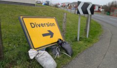 A yellow "Diversion" road sign with a right arrow is propped up on grass with sandbags, next to a road curve sign by a roadside fence.