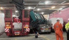A truck tilted and crashed onto a car and van in a parking garage, while three workers in safety gear observe the scene.