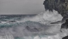 Waves crash against a rocky cliff under a cloudy, overcast sky, with a pebble-strewn shoreline in the foreground.