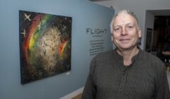 A man stands in front of a blue wall displaying a painting of birds in flight and a rainbow, with a sign reading "FLIGHT by Paul Bibrone.