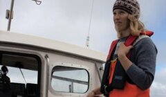 A woman wearing a knit hat and life jacket stands on a boat near a cabin window, looking off to the side.
