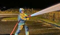 A firefighter in full gear sprays water from a hose in a parking lot at night.