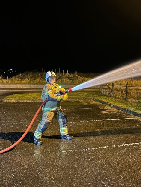A firefighter in full gear sprays water from a hose in a parking lot at night.