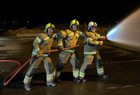 Three firefighters in full gear operate a hose, spraying water at night on a wet outdoor surface.