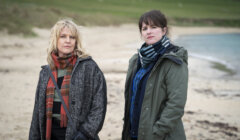 Two women wearing coats and scarves stand on a beach with sand and grass in the background, looking toward the camera.