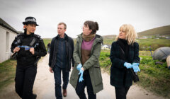 Four people, including a uniformed police officer and three others wearing gloves, walk and talk together along a rural path with fields and houses in the background.