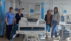 Four women stand around a hospital bed in a brightly lit hospital room, with medical equipment and chairs visible in the background.