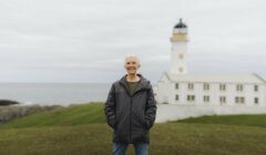 An older man stands outdoors with his hands in his jacket pockets; a white lighthouse and building are visible in the background near the sea.