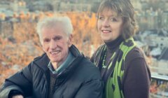 An older man and woman stand side by side outdoors, smiling at the camera with a cityscape and trees in the background during sunset.