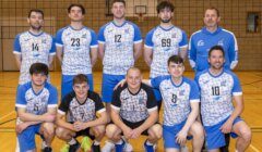 A men's volleyball team poses for a group photo in a gym, with five players kneeling in front and six standing behind. All wear blue and white uniforms.
