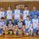 A men's volleyball team poses for a group photo in a gym, with five players kneeling in front and six standing behind. All wear blue and white uniforms.