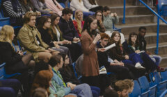 A woman stands and speaks into a microphone while reading from a sheet of paper in a seated audience at an indoor event.