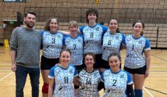A volleyball team of eight women and one man stands on an indoor court posing for a group photo; the players wear matching blue and white uniforms.