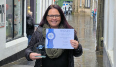 A woman stands in a wet alley holding a winner’s certificate and a glass award, smiling at the camera.