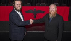 Two men in suits shake hands in front of a red banner featuring a black bird emblem.