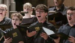 A group of choir singers, dressed in black and holding NYCoS folders, perform indoors with expressive faces.
