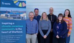 Six people stand in front of a light-colored wall next to a sign for the Brae Rural Energy Hub, which promotes climate-conscious community initiatives in Shetland.