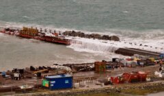 A construction site by the sea shows heavy machinery, building materials, and barriers with waves breaking over a partially built seawall.