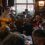 Two musicians perform with guitars in a crowded pub as people sit at tables and clap along, with stained glass windows in the background.