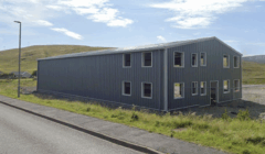 A large, grey, metal industrial building with multiple windows stands beside a paved road on a grassy landscape under a partly cloudy sky.