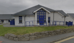 Single-story white building with a gray roof, large blue-framed windows and doors, a blue accessibility ramp, and a stone retaining wall in front.