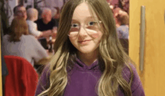 A girl with long, light brown hair and glasses, wearing a purple hoodie, stands indoors with people sitting and talking in the background.