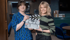 Two women stand indoors holding a clapperboard labeled "Shetland" in a room with office furniture and shelves in the background.