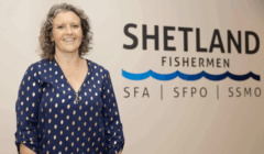 A woman stands smiling in front of a wall displaying the logo and text for Shetland Fishermen, SFA, SFPO, and SSMO.