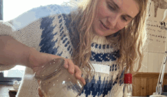 A woman in a patterned sweater pours liquid from a glass jar into a clear bottle at a wooden table indoors.
