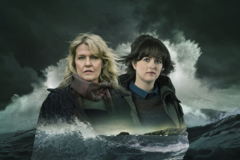 Two women stand in front of stormy ocean waves under a dark, cloudy sky.