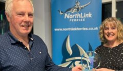 Two people stand indoors, smiling, as one hands an award to the other in front of a NorthLink Ferries banner.