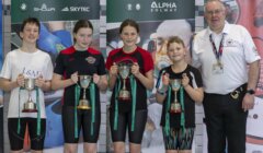 Four young swimmers holding trophies stand next to an adult in a white shirt and lanyard, posing for a group photo indoors with banners in the background.