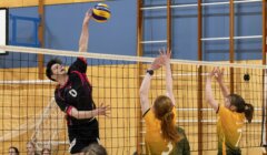 A volleyball player in a black uniform jumps to spike the ball over the net as two opponents in yellow attempt to block in an indoor gym.