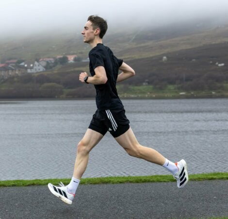A man in athletic clothing runs on a paved path beside a lake, with hills and fog in the background.