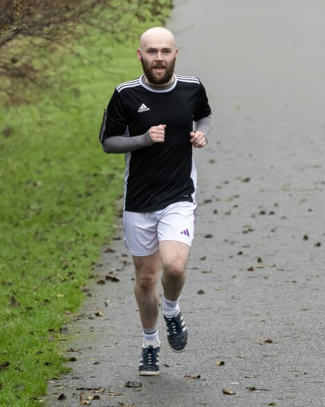 A man wearing a black shirt and white shorts is jogging on a paved path outdoors, with grass and scattered leaves nearby.