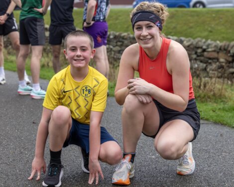 A smiling woman and boy in athletic clothing crouch on a path outdoors, with other runners and a stone wall in the background.