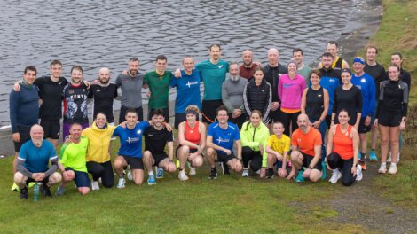 A group of about 30 people in athletic clothing pose together on grass beside a body of water, appearing to have completed an outdoor group activity.