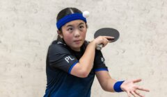 A young girl prepares to serve in a game of table tennis, holding a paddle and tossing a ball in the air.