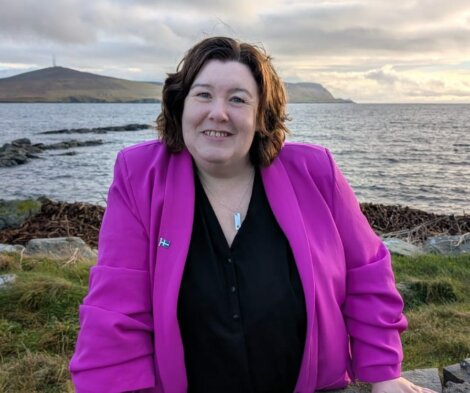 A woman wearing a bright pink blazer stands outdoors by the water with hills in the background, smiling at the camera.