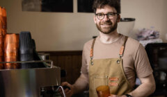 A man wearing glasses and an apron stands by an espresso machine, holding a brown cup and smiling at the camera in a coffee shop setting.