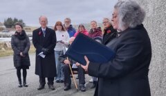 A group of people stand and sit outdoors near a stone wall as a woman in a dark coat speaks and holds a large book. Most people wear poppy pins. The weather appears cloudy and cold.