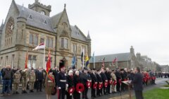 A formal outdoor ceremony with uniformed soldiers and civilians holding wreaths in front of a historic stone building, with flags displayed and a speaker at a podium.