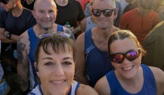 Group of runners in athletic gear pose for a selfie at a race event, surrounded by other participants, in early morning sunlight.