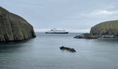 A ferry labeled "ORKNEY FERRY" sails between two rocky cliffs on calm water under a cloudy sky.