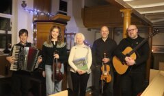 Five people stand indoors, three holding musical instruments (accordion, violin, guitar), one holding a book, and another standing with hands by their sides; string lights are visible in the background.