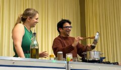 Two people stand behind a counter with cooking ingredients and utensils; one stirs a pot on a stove while the other smiles. Both wear head microphones.