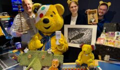 Four people, one in a Pudsey Bear costume, pose in a radio studio surrounded by various toys, crafts, and gift items, all supporting BBC Children in Need.