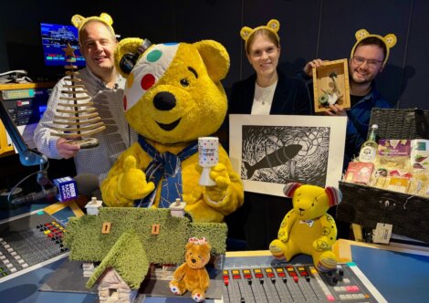 Four people, one in a Pudsey Bear costume, pose in a radio studio surrounded by various toys, crafts, and gift items, all supporting BBC Children in Need.