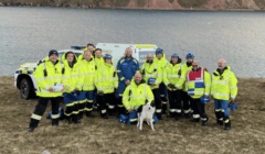 A group of rescue workers in yellow jackets and helmets stand on grass by a body of water, with a dog and emergency vehicles visible behind them.